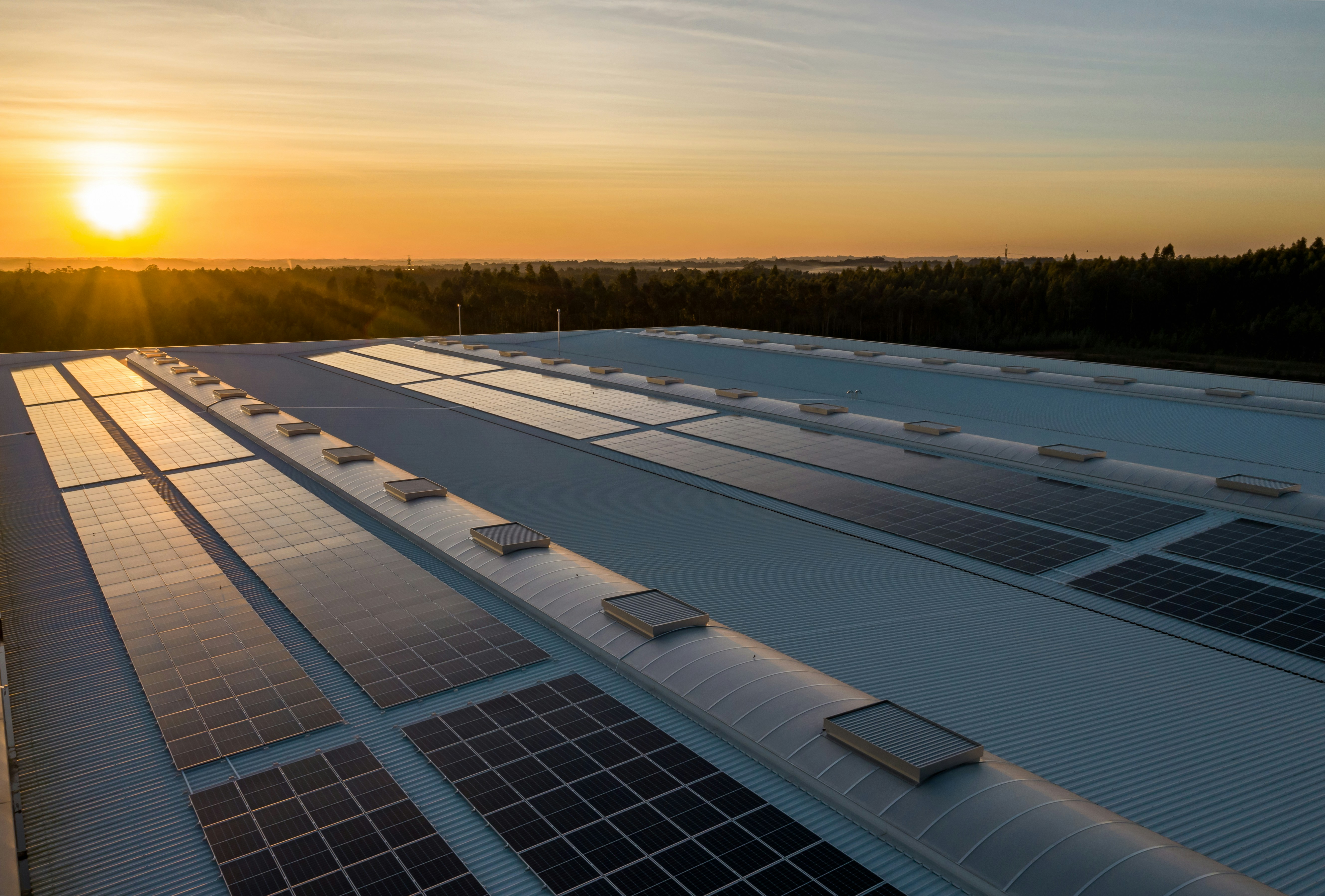 Solar panel arrays on the roof of a huge industrial building, with sun rising in the background